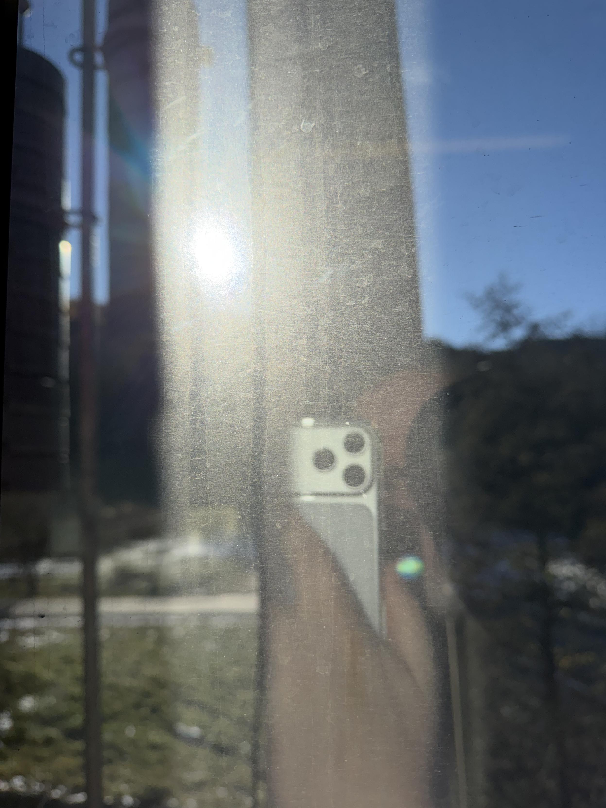Reflexionsaufnahme durch ein schmutziges Fenster, in dem ein heller Sonnenlichtfleck und das teilweise Spiegelbild einer Hand mit Smartphone zu sehen sind; draußen verschwommen eine sonnige Landschaft mit blauem Himmel und etwas Grün.