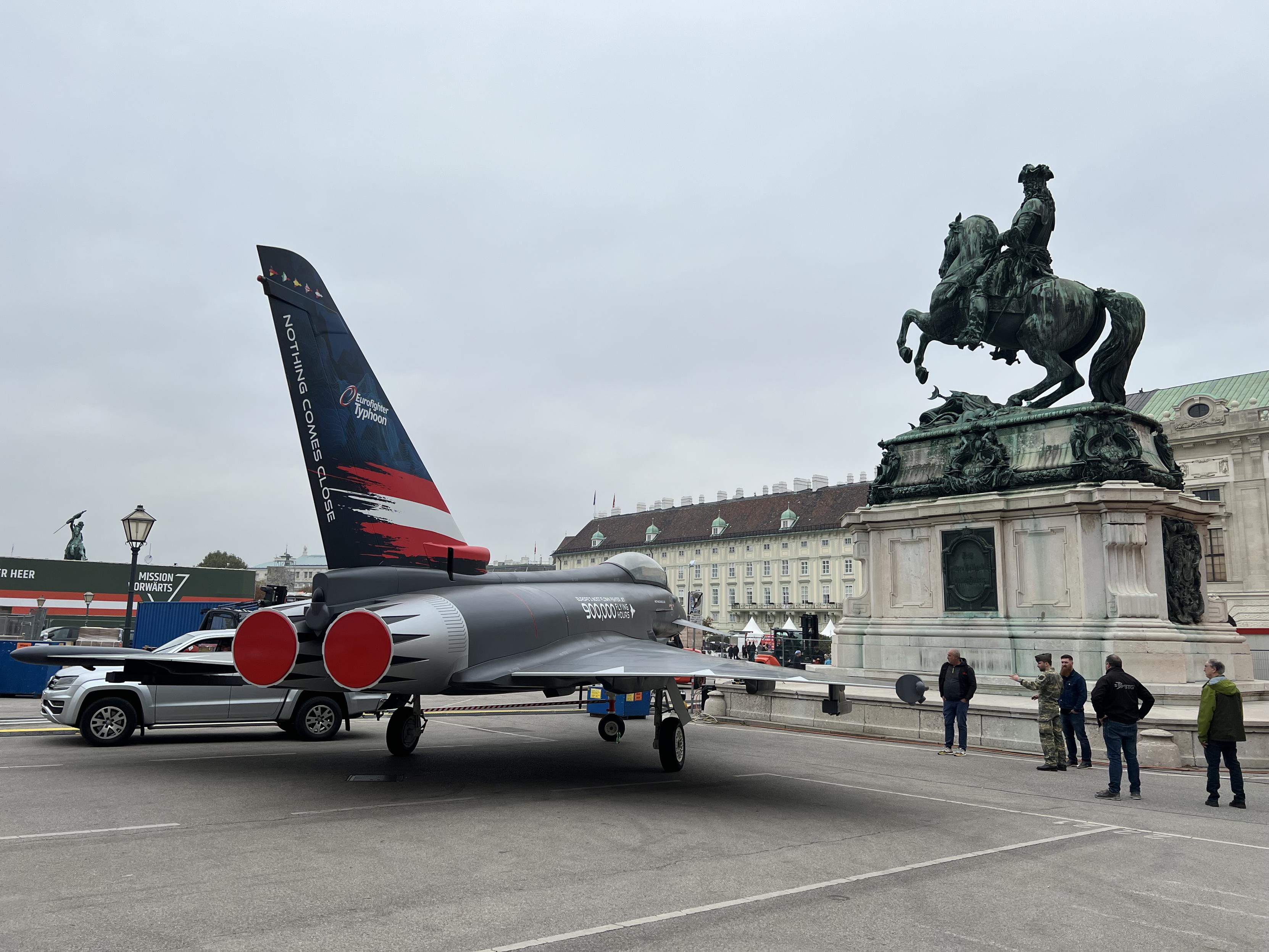 Das Bild zeigt einen Eurofighter Typhoo  mit der Aufschrift “Nothing Comes Close” auf der Heckflosse, der am Heldenplatz, neben der Reiterstatue von Prinz Eugen ausgestellt ist.