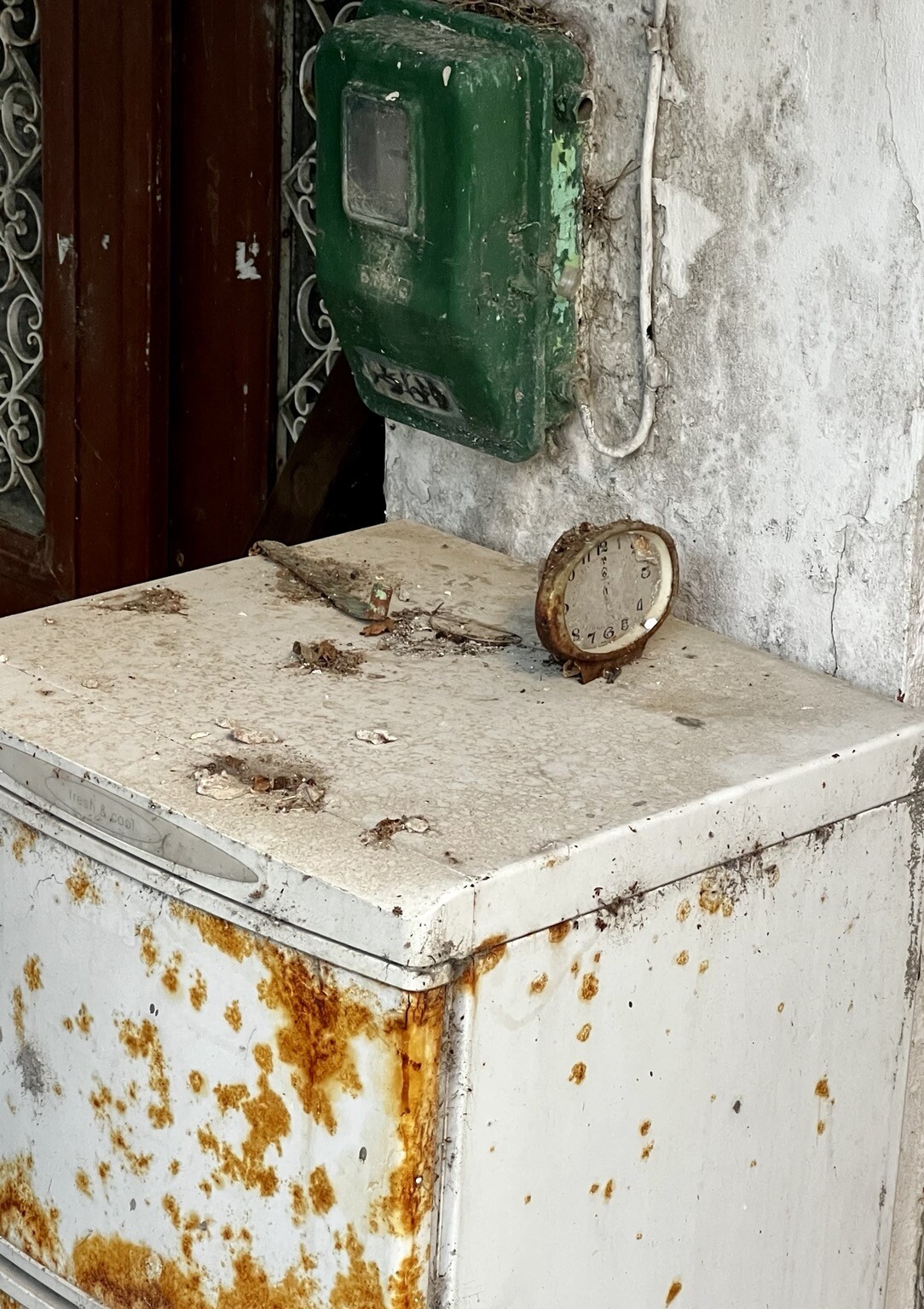 Verrosteter Wecker ohne Zeiger steht auf verrosten Kühlschrank. An der Wand hängt ein alter Zählerkasten