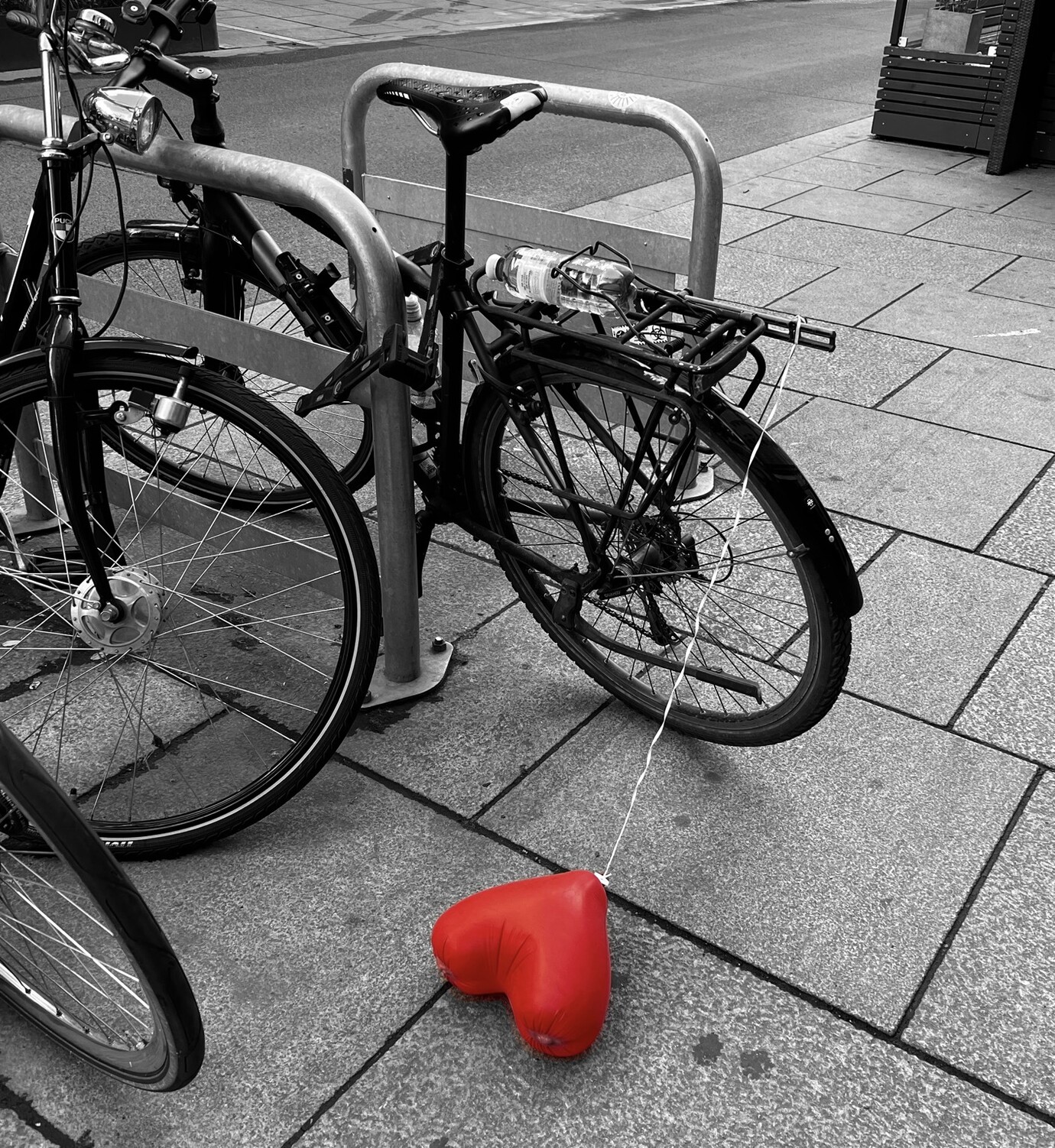 Fahrräder an einem Metallrahmen gelehnt. An einem Rad hängt eine Schnur. Diese führt runter zum Gehsteig auf dem ein roter herzförmiger Luftballon liegt.