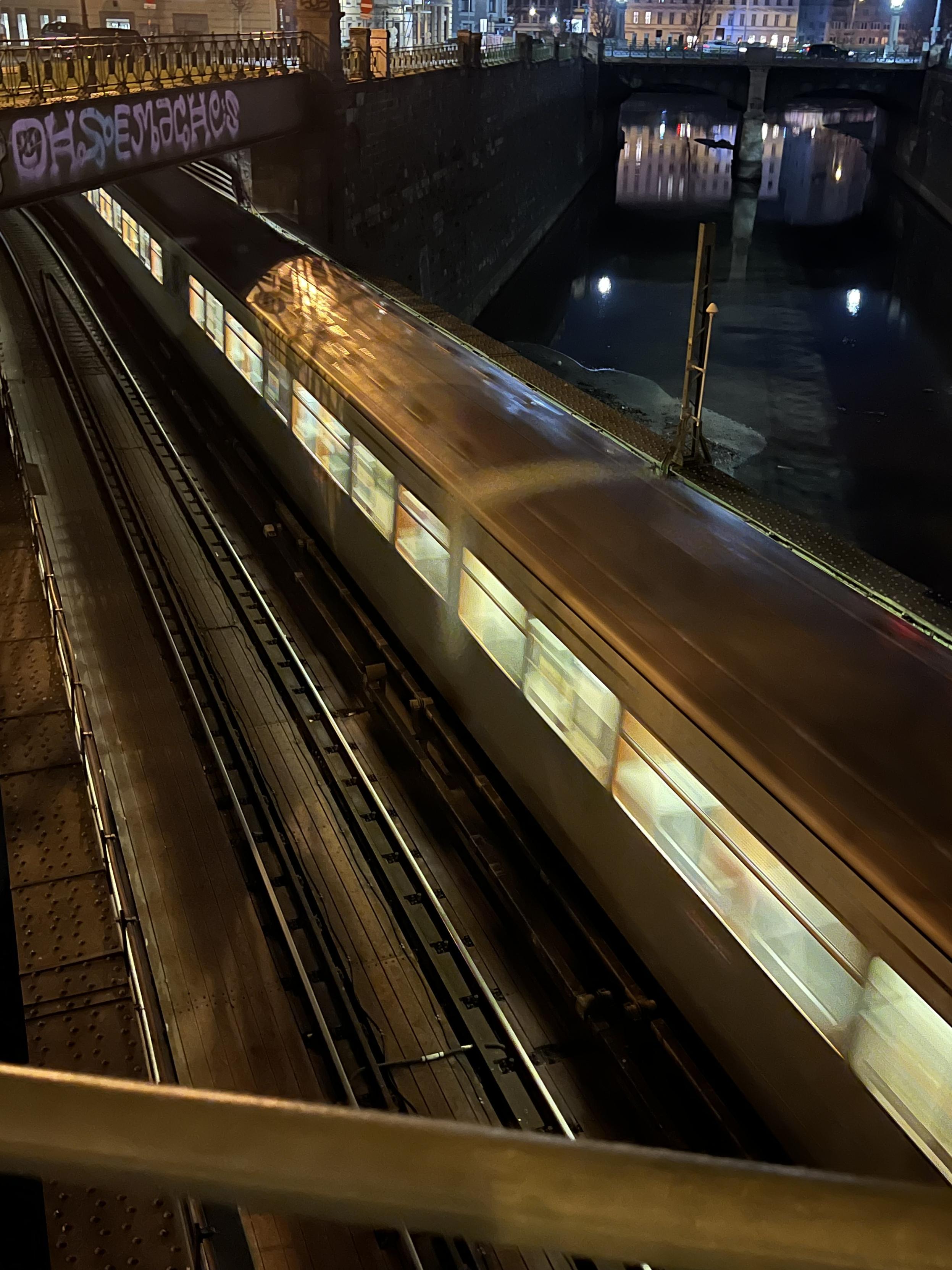 Ein nächtlicher Blick auf eine vorbeifahrende U-Bahn auf erhöhten Gleisen, unter einer Brücke mit Graffiti, flankiert von einem Kanal. Die Lichter der U-Bahn spiegeln sich auf den Schienen und der Umgebung wider.