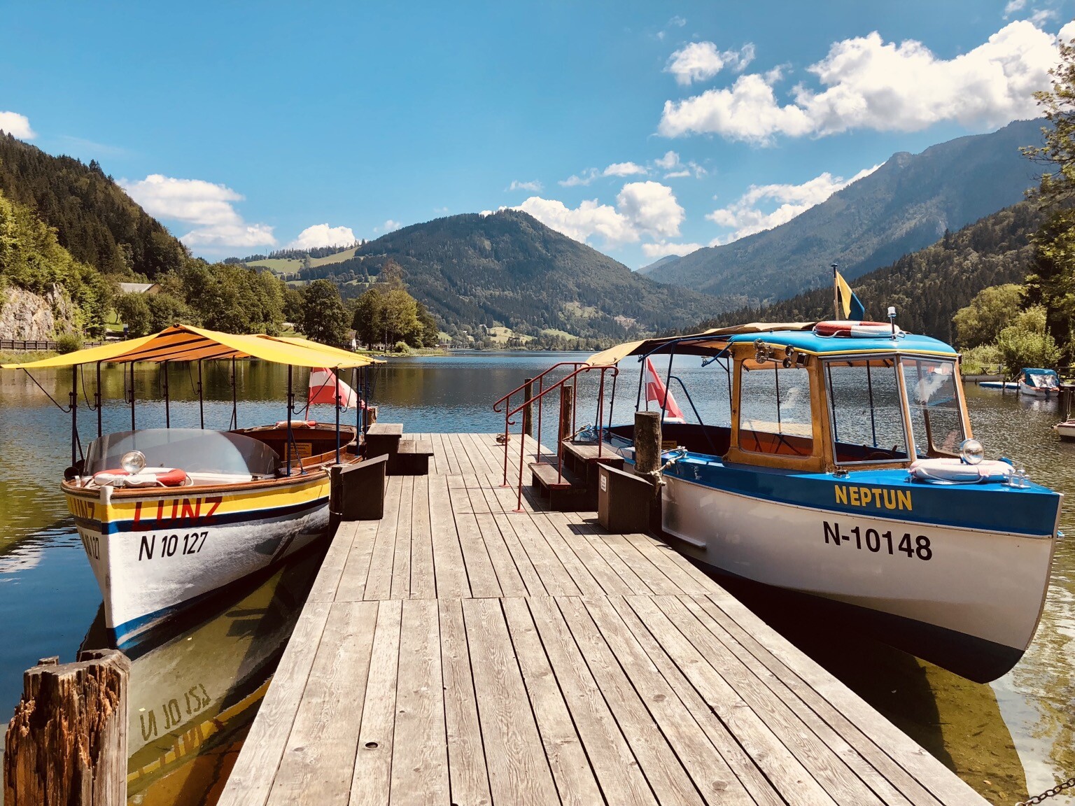 Blick auf See. Bootssteg mit zwei kleinen überdachten Ausflugsbooten. Dahinter begrünte Berge.